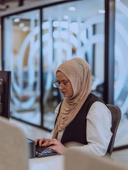 In a modern office, a young Muslim entrepreneur wearing a hijab sits confidently and diligently works on her computer, embodying determination, creativity, and empowerment in the business world