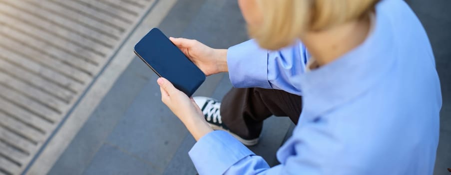 Close up of woman looking at smartphone, cropped shot of mobile phone in female hands, girl sitting on stairs and using telephone.