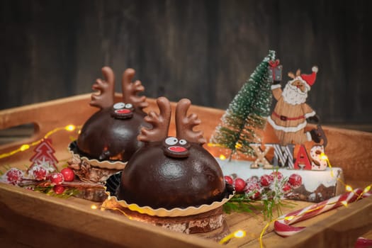 Two chocolate deer in a wooden tray with sweets, confetti, a burning garland and a christmas decoration on a wooden table, close-up side view.