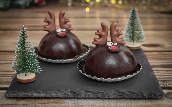 Two chocolate deer on a black stone board with Christmas trees on a brown wooden table, close-up side view.
