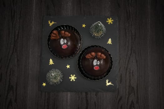 Two chocolate deer cakes on a black stone board with fir trees, christmas decoration and a burning garland on a black table, close-up side view.