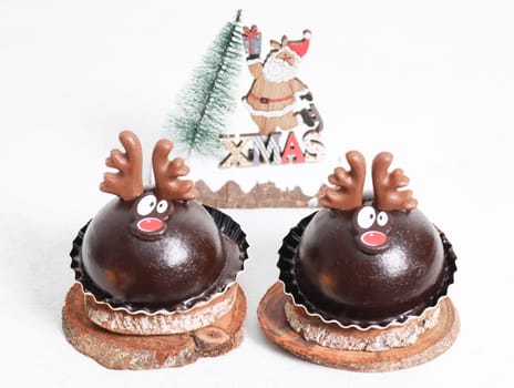 Two chocolate cake deer on wooden cuts lies on a white cement table with a blurred Christmas tree and santa claus on the background, close-up side view.