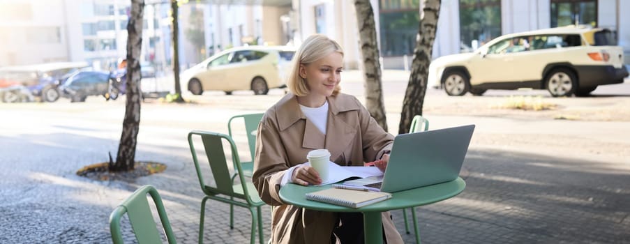 Image of young modern woman, sitting in cafe outdoors, drinking coffee or tea, looking at laptop, working on project, freelancing, making notes in notebook. Lifestyle concept