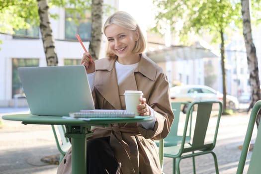 Image of young blond modern woman, sitting with laptop outside in cafe, drinking coffee drink and working on project, using computer.