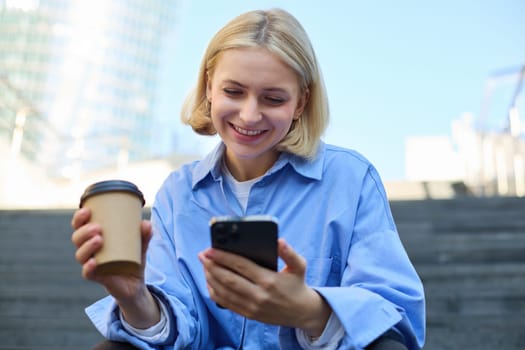 Close up portrait of young smiling woman with cup of coffee, drinking and sitting on stairs in city, holding smartphone in hand.