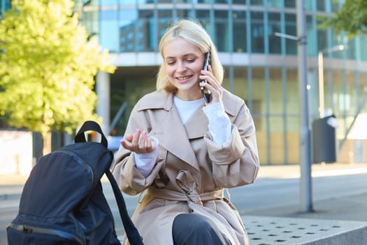 Portrait of young woman sitting on bench, talking on mobile phone, spending time outside, answers a telephone call, having friendly conversation on cellphone.