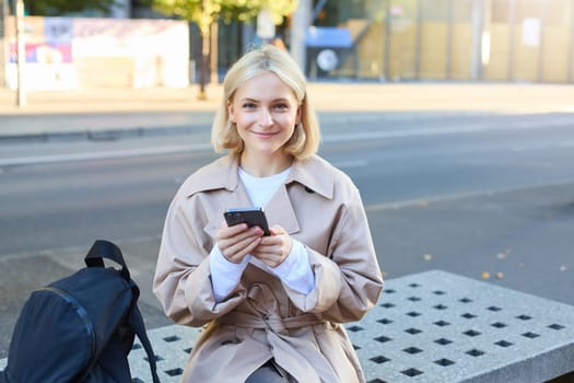 Outdoor shot of young blonde woman on street, sitting on bench with mobile phone, smiling at camera, waiting for friend outside of building, having a break.