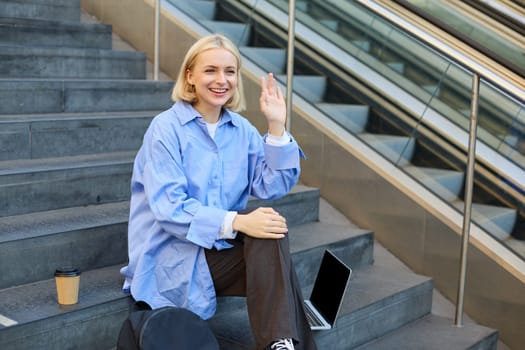 Friendly smiling woman, student waving hand to say hi to friend or classmate, sitting outdoors on campus stairs, resting after working on laptop, drinking coffee.