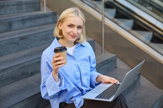 Image of stylish young modern woman, student doing homework, studying outdoors on campus stairs, sitting with laptop and coffee, drinking her cappuccino and connecting to public wifi.