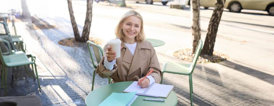 Smiling, cheerful woman drinking her coffee in street cafe, writing in notebook, doing homework, making notes. Student and lifestyle concept.