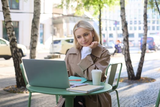 Image of young woman looking at laptop, sitting in outdoor cafe and drinking coffee, studying remotely, attends online course while sits outside, working, freelancing.