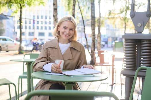 Portrait of young blonde woman sitting in cafe, doing homework and drinking coffee, writiing in journal, using notebook on fresh air.