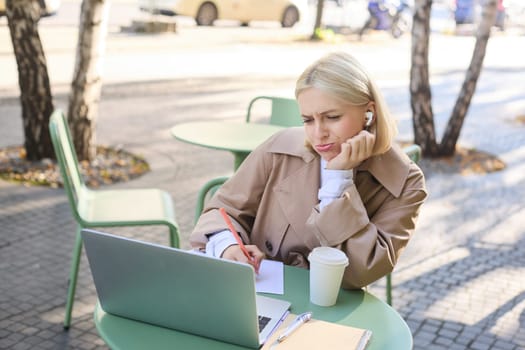 Portrait of blond girl with upset face, listening lecture, attends online course in wireless headphones, sits in outdoor cafe and frowns while makes notes, studying remotely, using laptop.