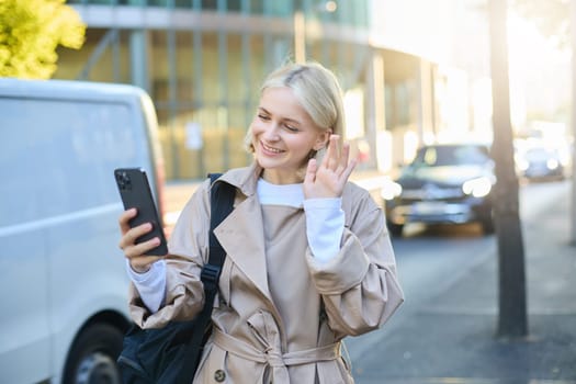 Portrait of happy young woman walking on street, waving hello at smartphone camera, connects to online video chat, calling someone on mobile phone on her way to work.