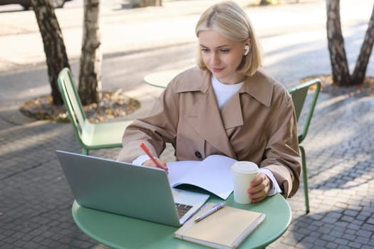 Portrait of beautiful blond woman, wearing wireless headphones, using laptop, studying in outdoor coffee shop, making notes, working on project.