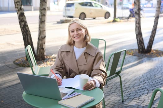 Portrait of young woman working on project, sitting outdoors in cafe, drinking her coffee, using laptop and making notes in notebook, studying, preparing for work interview.