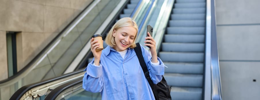 Image of young happy blond woman, dancing from happiness and joy, drinking takeaway coffee, holding smartphone, posing near escalator in city centre.