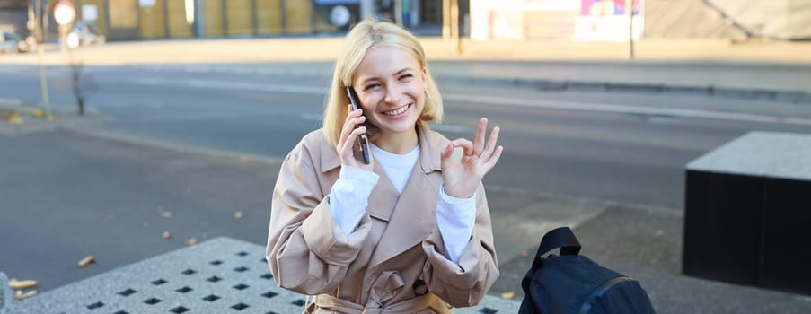 Happy beautiful young woman sitting on bench with mobile phone, talking on cellphone and showing okay gesture. Lifestyle and people concept