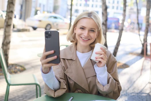 Beautiful young blond woman, posing with takeaway cup in outdoor cafe, sitting on street with coffee, taking selfie, recording live stream from favourite shop.
