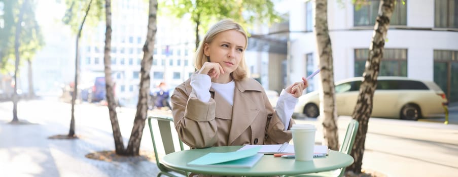 Thinking woman, student sitting in cafe with notebook, drinking coffee and doing homework, frowning while pondering.