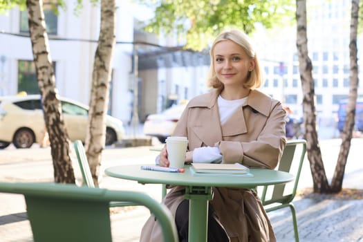 Image of young woman, makes sketches outdoors on street in notebook, drinks coffee and smiles at camera. Lifestyle and people concept