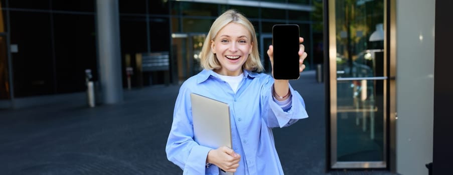 Happy smiling woman showing smartphone screen, demonstrating mobile phone display, standing in college outdoors, posing near campus with laptop and telephone.