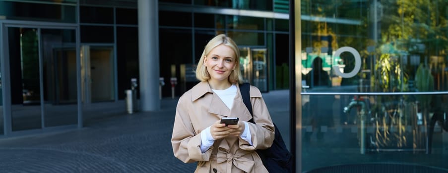 Street photo of young blond woman with smartphone, walking in city, waiting for someone, order taxi on mobile application.