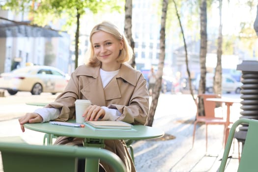 Portrait of young blonde woman sitting in cafe, doing homework and drinking coffee, writiing in journal, using notebook on fresh air.