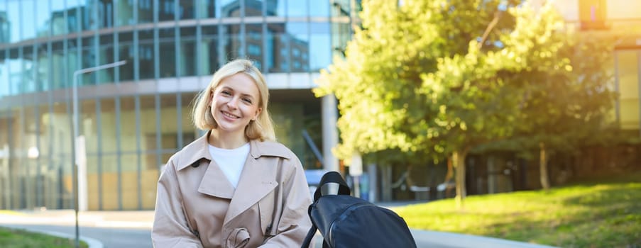 Vertical shot of cute blond woman in trench coat, sitting on street bench with smartphone, chatting with friend, waiting for someone outside, smiling and looking at camera.