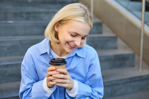 Close up portrait of beautiful, smiling blonde woman, student sitting on stairs outside campus, drinking takeaway coffee, warm-up her hands while holding a cup.