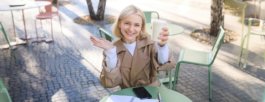 Food and drink concept. Young happy woman sitting in cafe, enjoying bright day outdoors, raising cup of coffee and smiling, drinking chai.