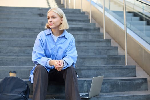 Urban style portrait of young woman, student sitting on city street stairs, with backpack, cup of coffee and laptop, looking happy and upbeat, smiling.