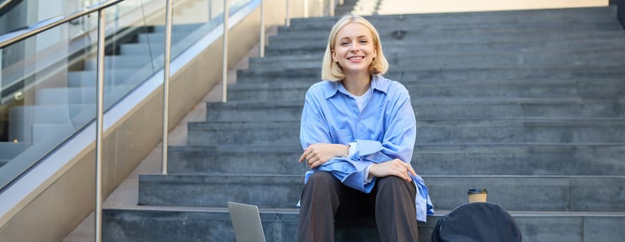 Close up portrait of young cheerful woman, looking confident at camera, sitting on street, urban stairs with laptop, backpack and cup of coffee.