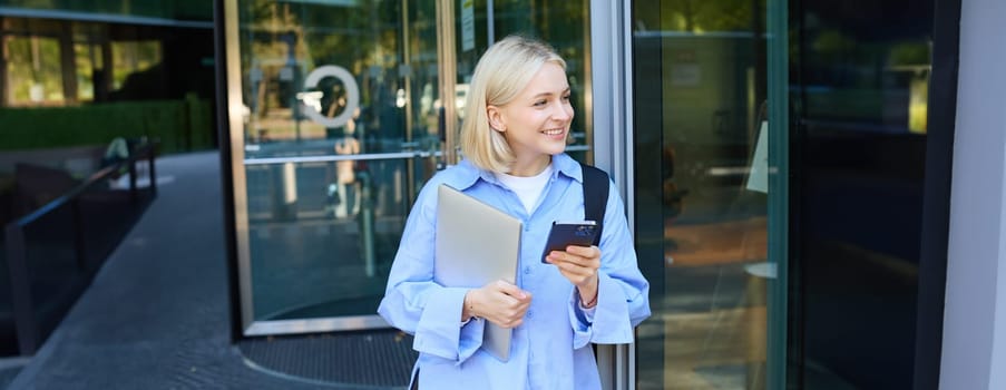 Image of stylish young woman, student walking out of the college campus, holding smartphone and laptop, has backpack on shoulder, waiting for someone outside with mobile phone.