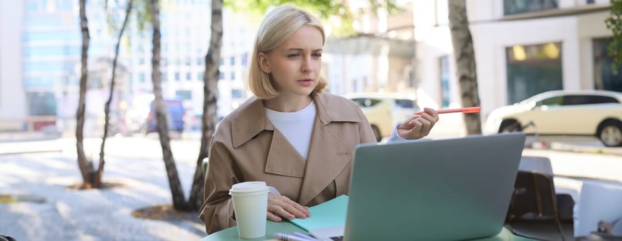 Portrait of young blond woman, modern female model sitting in outdoor cafe, drinking coffee, connects to online video chat via laptop, talking to coworkers remotely, gesturing and explaining smth.