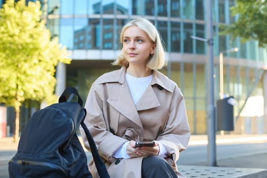 Portrait of young blond woman, student sitting on street bench, waiting for someone, holding mobile phone, looking around, order taxi on smartphone app.