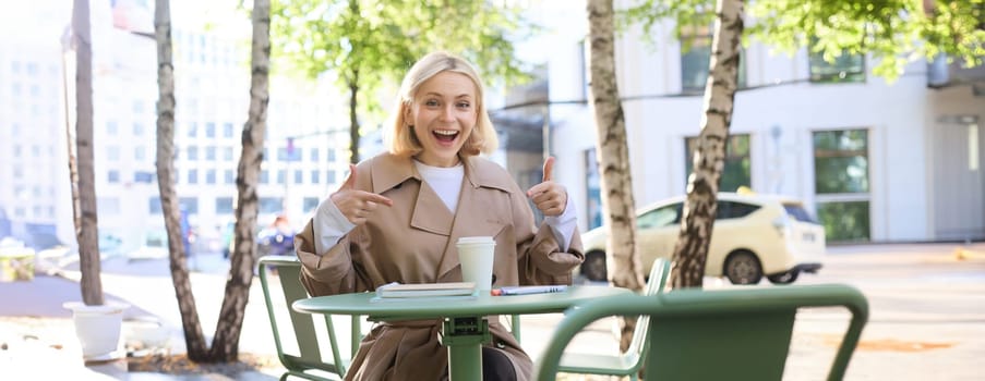 Portrait of excited beautiful girl, pointing fingers at takeaway cup, order awesome drink in cafe, recommending coffee place.