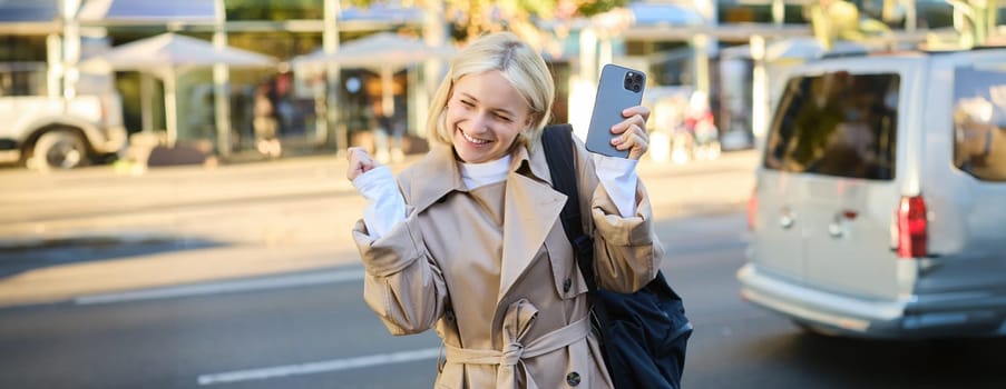 Cheerful young blond woman, holding smartphone, cheering and rejoicing on street, celebrating victory, raising hands up and triumphing, making fist pump gesture.