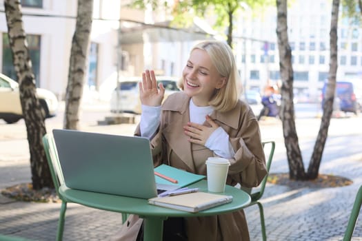 Portrait of young friendly woman, waving hand at laptop and smiling, saying hello, greeting someone, online chatting, having remote conversation while sitting in coffee shop.
