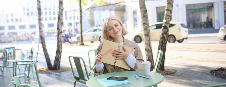 Image of young smiling blond woman sitting in an outdoor cafe, holding notebook, doing her homework outside in coffee shop, looking happy.