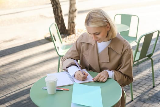 Portrait of woman working on project, writing in notebook, sitting outdoors in cafe, drinking coffee and doing homework. Student lifestyle and people concept
