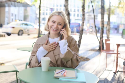 Happy blond female model, sitting outdoors in cafe, answer phone call, having conversation on smartphone, laughing and smiling. Lifestyle and people concept