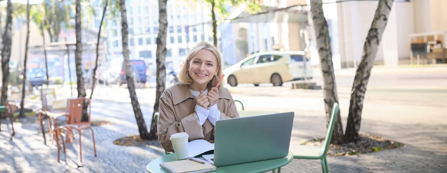 Portrait of excited blond woman, student in outdoor cafe, sitting in coffee shop with laptop, looking happy and amused, holding pen with notebook, studying or doing homework.
