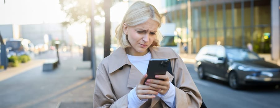 Portrait of young female model on street, looking at smartphone with disappointed, upset face, frowning while reading mobile phone message.