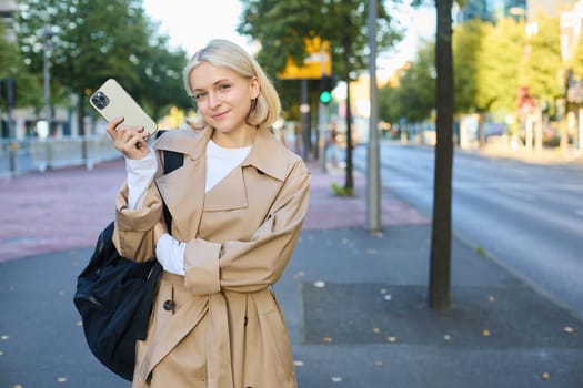 Image of young modern woman in trench coat, student holding mobile phone and backpack, posing on streets, standing near road, smiling and looking at camera.
