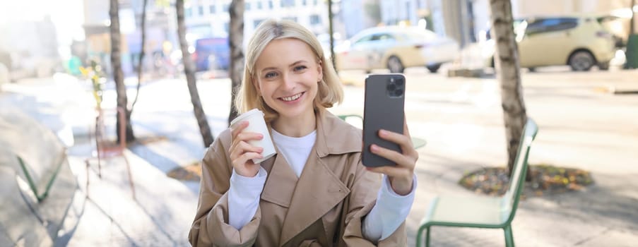 Beautiful young blond woman, posing with takeaway cup in outdoor cafe, sitting on street with coffee, taking selfie, recording live stream from favourite shop.