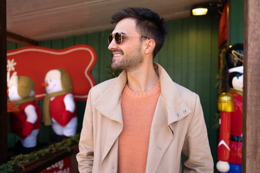 Stylish man in sunglasses smiles looking side standing outdoor. Bearded male standing near Christmas decoration outside. Happy people positive emotions