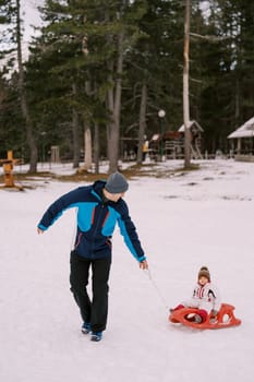 Dad carries a small child on a sleigh along the snowy edge of the forest, looking back. High quality photo