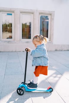 Little girl rides a three-wheeled scooter holding the steering wheel and looking down at her feet. Side view. High quality photo