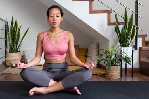 Young Asian woman meditating at home sitting on yoga mat. Spirituality and mindfulness concept.